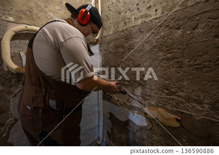 A man uses a hammer drill to remove old tiles from the walls. A construction worker, wearing ear protection and a respirator, uses a hammer drill to remove old plaster and cement from the wall. A man uses a hammer drill to remove old tiles from the walls. A construction worker, wearing ear protection and a respirator, uses a hammer drill to remove old plaster and cement from the wall. 136590886
