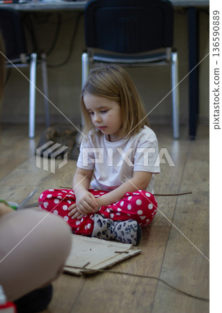 Vertical. A girl sits on a wooden floor, intently creating crafts from natural materials and cardboard during an art workshop. The child develops creativity through learning. 136590889