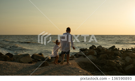 Children stroll along the rocky coastline, holding hands. They gaze out at the endless ocean under the warm evening sky, enjoying the feeling of togetherness during the holidays. Children stroll along the rocky coastline, holding hands. They gaze out at the endless ocean under the warm evening sky, enjoying the feeling of togetherness during the holidays. 136590893