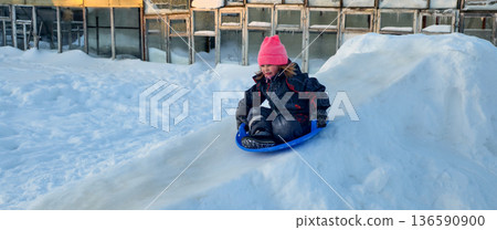 A child sleds down a snowy hill, wearing a pink hat and winter clothes. The child enjoys the thrill and fun of outdoor winter activities and relaxing in cold weather under natural light. 136590900