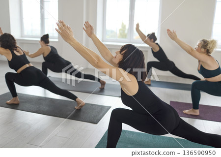 Focused women's yoga group holding high lunge pose during class in sunlit studio Focused women's yoga group holding high lunge pose during class in sunlit studio 136590950