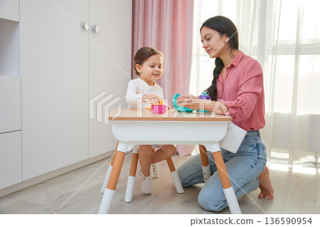 Mother and daughter enjoying a pretend tea party with colorful toy dishes 136590954