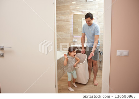 Father and daughter placing dirty clothes into a laundry hamper at home 136590963