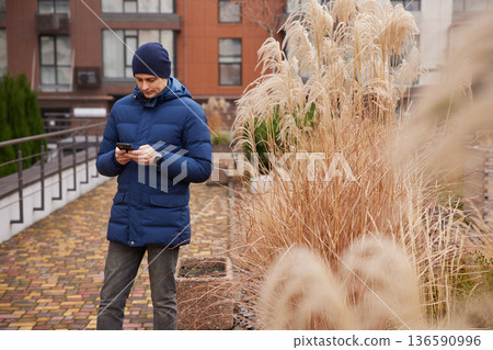 Man in winter coat and beanie texting on phone outdoors near ornamental grass 136590996
