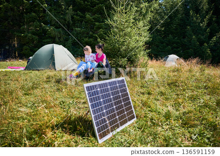 Young child and mother playing near tourist tent with photovoltaic solar panel in summer. On background of dense forest. Integration of renewable energy in outdoor camping activities. Young child and mother playing near tourist tent with photovoltaic solar panel in summer. On background of dense forest. Integration of renewable energy in outdoor camping activities. 136591159