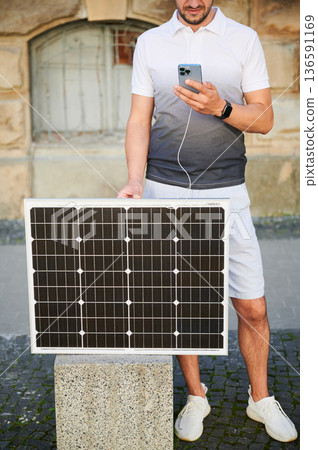 Cropped photo of man charging phone by portable solar panel. Young male using solar battery charger for mobile devices. Concept of alternative energy resources and sustainable lifestyle. Cropped photo of man charging phone by portable solar panel. Young male using solar battery charger for mobile devices. Concept of alternative energy resources and sustainable lifestyle. 136591169