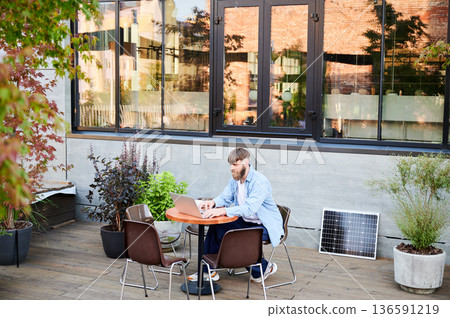 Bearded man works on laptop at outdoor round table, surrounded by greenery. Solar panel nearby, emphasizing sustainable, modern eco-friendly workspace that combines technology and sustainability Bearded man works on laptop at outdoor round table, surrounded by greenery. Solar panel nearby, emphasizing sustainable, modern eco-friendly workspace that combines technology and sustainability 136591219