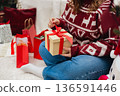 Merry Christmas, Happy Holidays banner. Cheerful pretty young woman having got a Christmas presents and sit on the floor near sofa and decorated Christmas tree. Cropped image 136591446