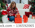 Merry Christmas, Happy Holidays banner. Cheerful pretty young 30s woman in red sweater having got a Christmas presents and sit on the floor near sofa and decorated Christmas tree. Full body 136592065