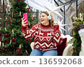 Cute young smiling woman listening music from smartphone during cozy Xmas holidays at home. Smiling Caucasian millennial lady in headphones makes selfie on phone in a Christmas decor 136592068