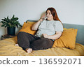 30s Woman on bed drinking a glass of water at home, relaxed and comfortable in bedroom, casual outfit, leisure moment, wellness and hydration concept 136592071
