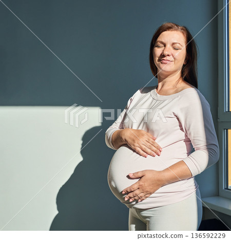 Pregnant caucasian female standing near window in sunlight holding belly Pregnant caucasian female standing near window in sunlight holding belly 136592229