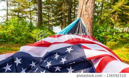 American flag draped over a hammock in a forest setting, surrounded by lush greenery and tall trees on a sunny day 136592310