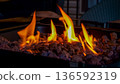 Flames dancing above black lava rocks in a modern fire pit, with blurred outdoor furniture in the background creating a cozy atmosphere 136592319