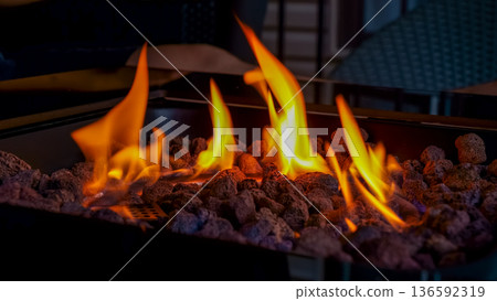 Flames dancing above black lava rocks in a modern fire pit, with blurred outdoor furniture in the background creating a cozy atmosphere Flames dancing above black lava rocks in a modern fire pit, with blurred outdoor furniture in the background creating a cozy atmosphere 136592319