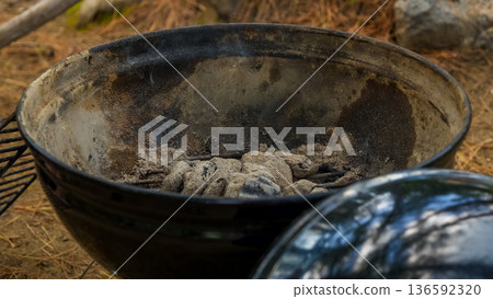 Charcoal grill with ashes and charcoal remnants, surrounded by natural outdoor setting with pine needles and rocks in the background 136592320
