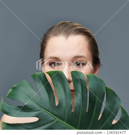 Caucasian female adult hiding behind monstera leaf against gray background Caucasian female adult hiding behind monstera leaf against gray background 136592477