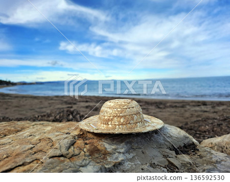 A straw hat sits on a rock at the beach. The sea is visible in the background along with some clouds. The sun shines brightly on the scene. A straw hat sits on a rock at the beach. The sea is visible in the background along with some clouds. The sun shines brightly on the scene. 136592530