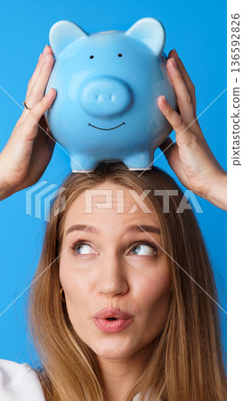 Young woman holds blue piggy bank above her head while looking up with a smile in studio setting Young woman holds blue piggy bank above her head while looking up with a smile in studio setting 136592826