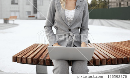 Woman works on laptop while seated on a bench in a public park during daylight hours Woman works on laptop while seated on a bench in a public park during daylight hours 136592876