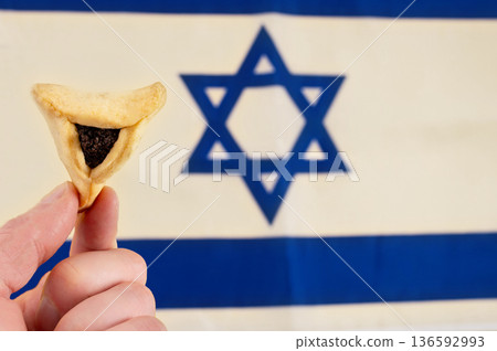 Traditional triangular cookie Hamantaschen in the hands on Israel flag background. Traditional food for Jewish holiday Purim. Traditional triangular cookie Hamantaschen in the hands on Israel flag background. Traditional food for Jewish holiday Purim. 136592993