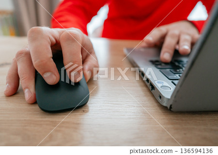 Close-up of a mans hand clicking a computer mouse while the other hand types on a laptop keyboard. Office or home workspace scene. Concept of remote work, productivity, online communication and Close-up of a mans hand clicking a computer mouse while the other hand types on a laptop keyboard. Office or home workspace scene. Concept of remote work, productivity, online communication and 136594136