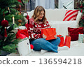 Merry Christmas, Happy Holidays banner. Cheerful pretty young 30s woman in red sweater having got a Christmas presents and sit on the floor near sofa and decorated Christmas tree. Full body 136594218