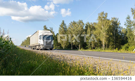 White semi-truck with a cargo trailer moves along an asphalt road on forest background. The truck drives along an highway White semi-truck with a cargo trailer moves along an asphalt road on forest background. The truck drives along an highway 136594416