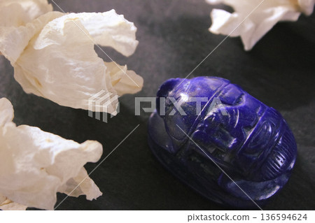 Lapis lazuli scarab beetle carving with dried white flowers on dark stone background, ancient symbol. Lapis lazuli scarab beetle carving with dried white flowers on dark stone background, ancient symbol. 136594624