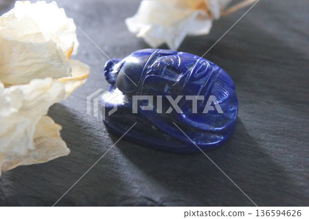 Lapis lazuli scarab beetle carving with dried white flowers on dark stone background, ancient symbol. 136594626