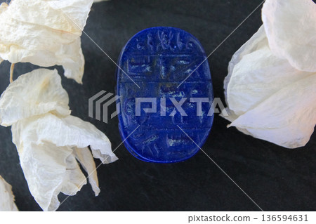 Lapis lazuli scarab base with engraved ancient egyptian hieroglyphs on dark stone with white petals. 136594631