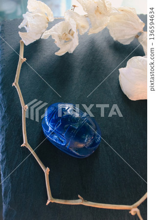 Lapis lazuli scarab beetle on dark slate surrounded by white petals and dry branches, sacred symbol. 136594634