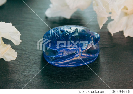Lapis lazuli scarab figurine on dark stone with white petals, ancient Egyptian protection symbol. Lapis lazuli scarab figurine on dark stone with white petals, ancient Egyptian protection symbol. 136594636