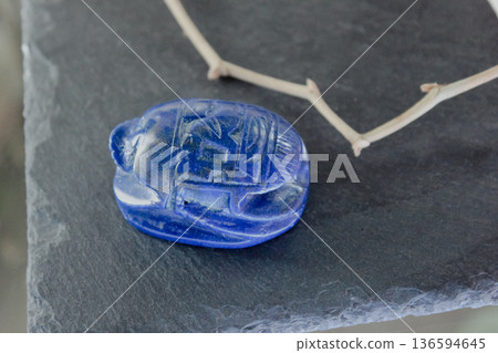 Lapis lazuli scarab beetle carving on dark slate stone with decorative dry branches, ancient amulet. Lapis lazuli scarab beetle carving on dark slate stone with decorative dry branches, ancient amulet. 136594645
