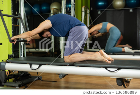 Young man performing pilates reformer spine stretch during group class Young man performing pilates reformer spine stretch during group class 136596899