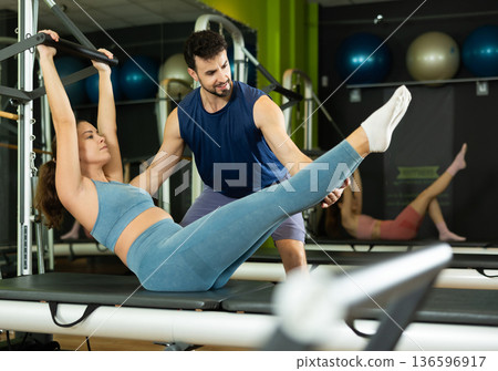 Woman doing abdominal exercises with the help of a male trainer and a reformer Woman doing abdominal exercises with the help of a male trainer and a reformer 136596917
