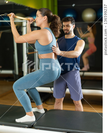 Male trainer guiding girl performing squat exercise on pilates reformer Male trainer guiding girl performing squat exercise on pilates reformer 136599237