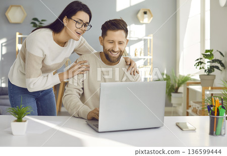 Happy loving woman supporting her husband who is working on his laptop at his desk 136599444