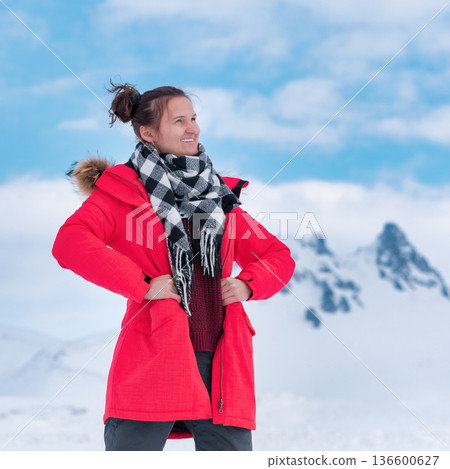 Joyful woman in bright red coat plaid scarf smiles against backdrop of snowy mountains blue sky. Natural daylight lighting. Concept of winter travel, happiness, outdoor adventure Joyful woman in bright red coat plaid scarf smiles against backdrop of snowy mountains blue sky. Natural daylight lighting. Concept of winter travel, happiness, outdoor adventure 136600627