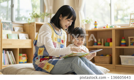 A nursery teacher sits on a cushion in the classroom and reads a picture book to children 136605643