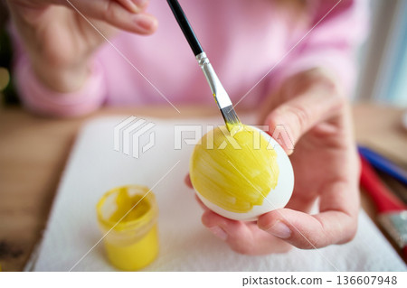 Close up of woman hands holding white egg while painting it with yellow paint and brush on table. Easter preparation and egg decorating for holiday concept Close up of woman hands holding white egg while painting it with yellow paint and brush on table. Easter preparation and egg decorating for holiday concept 136607948