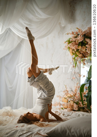 Woman performs yoga pose indoors using a bed and flowers in the background during daytime Woman performs yoga pose indoors using a bed and flowers in the background during daytime 136610610