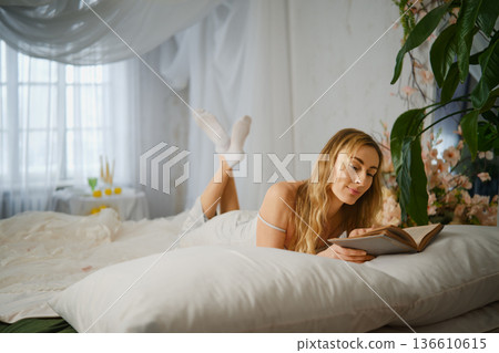Woman reads a book while lying on a bed in a bright room during the day with plants around Woman reads a book while lying on a bed in a bright room during the day with plants around 136610615