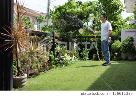A man waters plants in his sunny backyard with a relaxed smile. The morning light and green leaves express freshness and peaceful living. 136611383