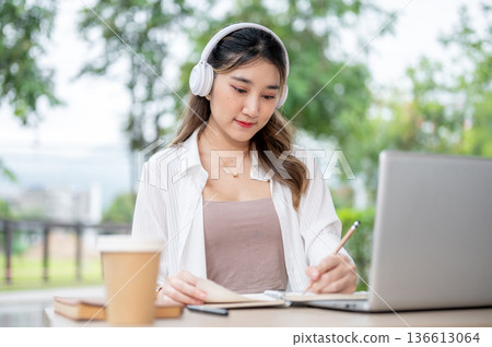 Young asian woman with headphone holding pencil writing in notebook in front of laptop on park table Young asian woman with headphone holding pencil writing in notebook in front of laptop on park table 136613064