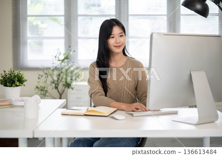 Pretty asian woman typing on computer keyboard aside notebook while sitting at white table in office Pretty asian woman typing on computer keyboard aside notebook while sitting at white table in office 136613484