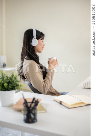 Asian woman wearings headphones holding coffee mug cup while sitting at office table or living room. Asian woman wearings headphones holding coffee mug cup while sitting at office table or living room. 136613508