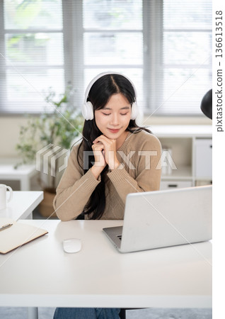 Asian woman wear headphones clasping hand under chin closing eyes in front of laptop on office table 136613518