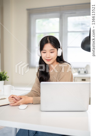 Asian woman wearing headphones looking at laptop on table while sitting in office or living room. Asian woman wearing headphones looking at laptop on table while sitting in office or living room. 136613521