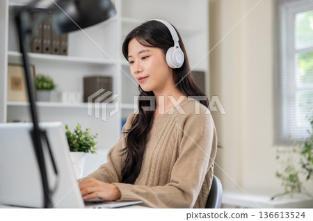 Asian woman wear headphones looking or typing on laptop while sitting at office table or living room 136613524